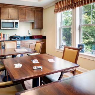 A cozy dining area with wooden tables and chairs, adjacent to a kitchen with wood cabinets and a microwave under natural light from large windows.