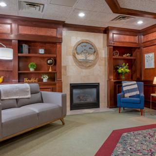 A cozy living room with wood paneling, featuring a fireplace, sofa, armchair, and built-in bookshelves.