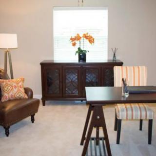 A cozy living room setup featuring a desk, chair, and a brown armchair with an elegant lamp. A decorative cabinet with an orchid adds a touch of style to the space.