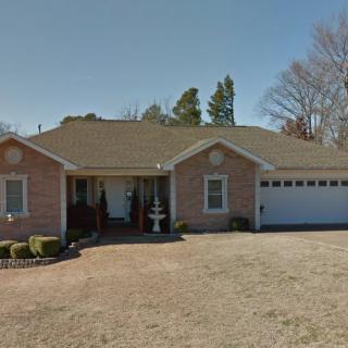 A single-story suburban house with a garage and a front lawn, bordered by trees under a clear sky.