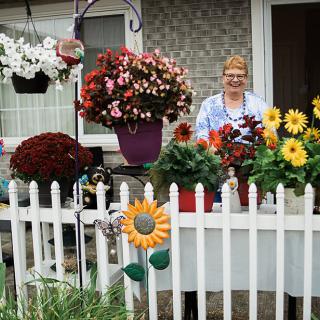 A woman stands behind a white fence surrounded by a variety of colorful flowers and potted plants.