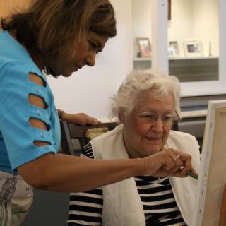 An elderly woman painting on a canvas with guidance from a teacher.