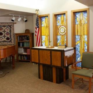 A chapel interior featuring stained glass windows, an altar, chairs, an American flag, and a piano.