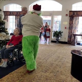 A festive outing as a person in a Grinch costume spreads holiday cheer to seniors in a community lobby.