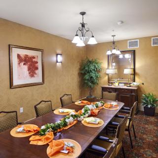 A well-decorated dining room with a long table set for a meal, featuring orange napkins and greenery centerpiece.