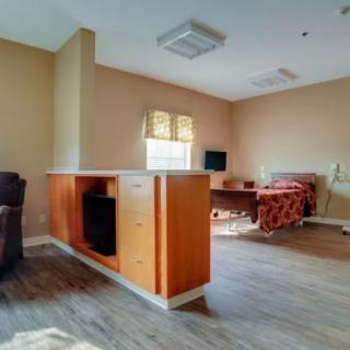 A cozy room with a bed, desk, and armchair, featuring wooden flooring and natural light from a window.