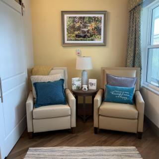 A cozy living room corner featuring two beige chairs with blue cushions, a small table with a lamp, and artwork on the wall by the window.