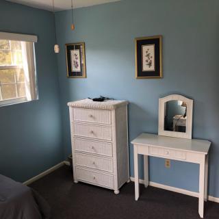 A cozy bedroom with blue walls, featuring a white dresser and vanity table, complemented by framed wall art.