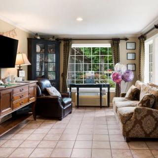 A cozy living room with tiled flooring, featuring a sofa, armchair, and a TV. Large windows allow natural light to illuminate the space.