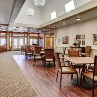 A spacious dining area with wooden flooring, tables, and chairs, illuminated by natural and ceiling lighting.