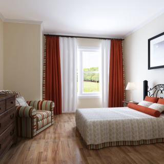 A cozy bedroom with a bed, dresser, and chair alongside a window with red and white curtains.