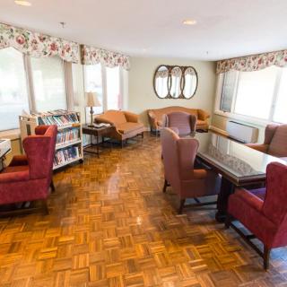 A cozy lounge area with comfortable seating, a table, and a bookshelf, featuring hardwood floors and large windows.