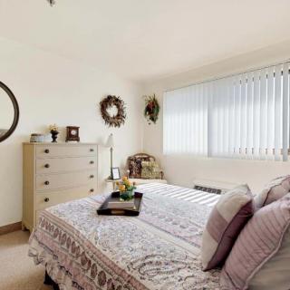 A cozy bedroom with a neatly made bed, adorned with pillows and a decorative tray. A dresser with a round mirror and framed photos enhances the warm atmosphere.