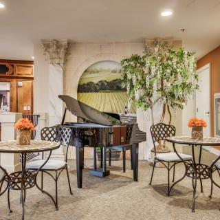 A cozy indoor setting featuring a grand piano surrounded by small tables and chairs, with decorative plants and artwork adorning the space.
