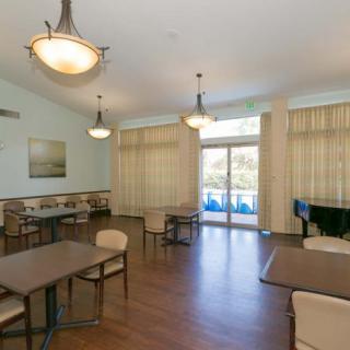 A dining area with tables, chairs, and a piano, featuring wooden floors and large windows with curtains.