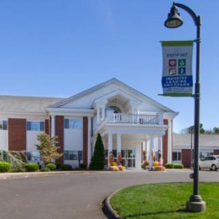 A large building with a grand entrance and white pillars, surrounded by greenery and featuring a banner on a nearby lamp post.