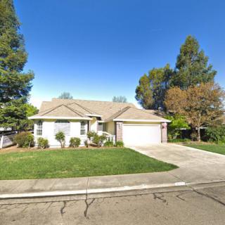A single-story suburban house with a well-maintained lawn and driveway, surrounded by trees under a clear blue sky.