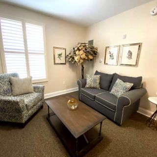 A cozy living room with a sofa, armchair, and coffee table, decorated with a potted plant and framed art on the walls.
