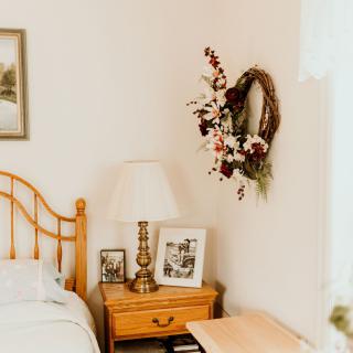 A cozy bedroom corner featuring a wooden nightstand with a lamp and photo frames, adorned with a floral wreath on the wall.