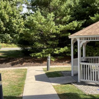 A sunny outdoor scene featuring a gazebo, surrounded by trees and greenery, with a pathway leading to it.
