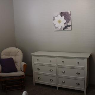 A minimalistic room featuring a white dresser, an armchair with a purple pillow, and a floral painting on the wall.