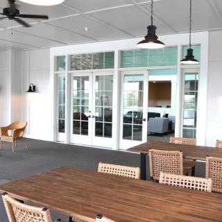 A modern indoor dining area with wooden tables and chairs, featuring pendant lights and a ceiling fan, leading to a room through glass doors.