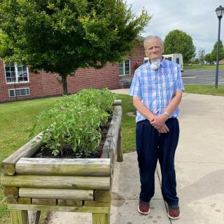 An elderly man standing next to a raised garden bed in an outdoor setting.