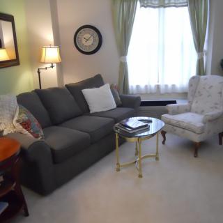 A cozy living room with a gray sofa, floral armchair, and round coffee table, illuminated by a floor lamp near a window with light curtains.