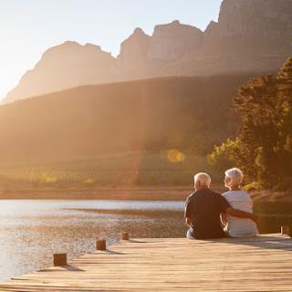 An elderly couple enjoys a serene sunset by the lake, surrounded by majestic mountains.