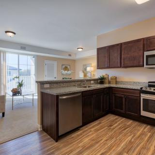 A modern kitchen with stainless steel appliances and wood flooring, connected to a bright living area.
