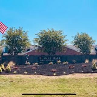 A landscaped area with a sign, surrounded by trees and an American flag in front of a brick building.