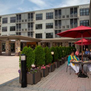 People enjoy outdoor dining on a patio with red umbrellas in an urban setting.