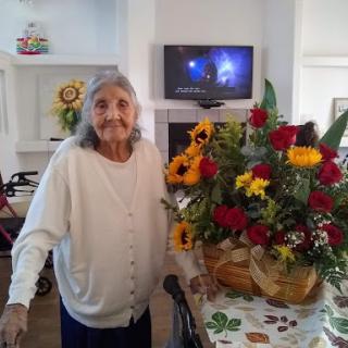 An elderly woman smiles proudly beside a vibrant bouquet of flowers in a warm, inviting setting.
