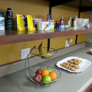 A kitchen counter displaying a fruit bowl with bananas, apples, and oranges, alongside a plate of cookies and various products on the shelf above.