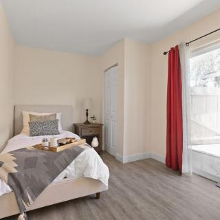A cozy bedroom with a single bed, natural light streaming in through red curtains, and a small nightstand.