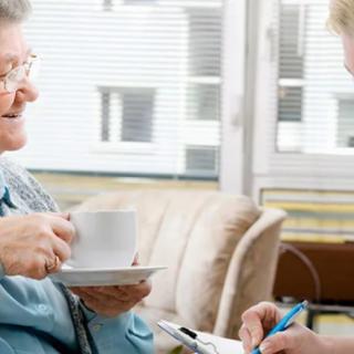 A caregiver visits an elderly person at home, discussing notes over a cup of tea.