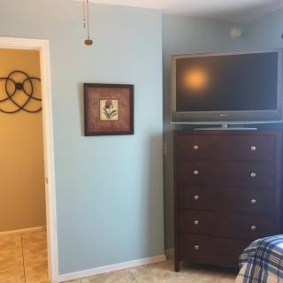 A bedroom with light blue walls featuring a wall-mounted TV above a tall dresser, adjacent to an open doorway and decorative wall art.
