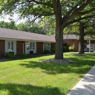 A view of a single-story building with a neatly maintained lawn, large trees, and a sidewalk on a sunny day.
