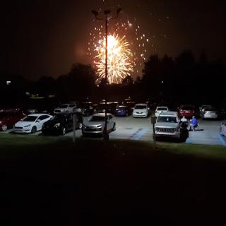 A stunning display of fireworks illuminates the night sky over a crowded parking lot.