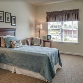 A cozy bedroom with a single bed, a chair by the window, and soft lighting from a floor lamp.