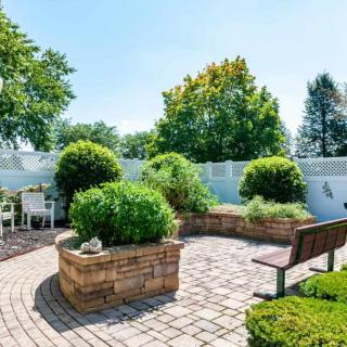 A beautifully landscaped garden with brick planters, lush greenery, and a wooden bench, enclosed by a white fence.