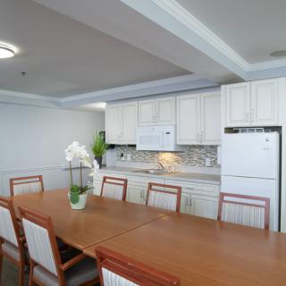 A modern dining area with a large wooden table, surrounded by chairs, adjacent to a white cabinet kitchen.