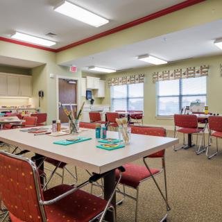 A bright and well-lit dining area with tables and chairs arranged neatly, featuring a small kitchen section.