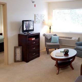 A cozy living room with modern furniture, including chairs and a round table, a television on a cabinet, and large windows allowing natural light.