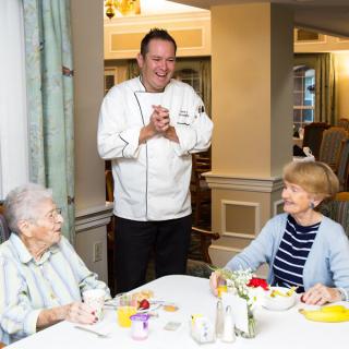 A chef interacting with two elderly women at a dining table.