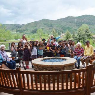 A large group of children and adults gather around a circular bench with a fire pit, set against a scenic mountain backdrop.