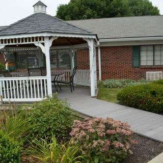 A peaceful garden gazebo with a bench, surrounded by lush greenery and adjoining a brick building.
