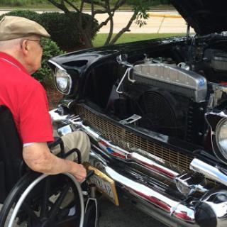 An elderly man in a wheelchair inspects the engine of a classic car.
