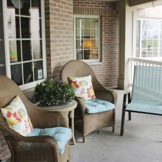 A cozy porch with wicker chairs, a small table, cushions, and a potted plant.