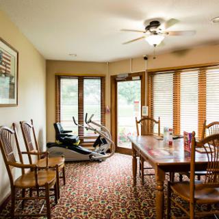 A cozy dining area featuring wooden furniture and an exercise machine, bathed in natural light from the large windows.